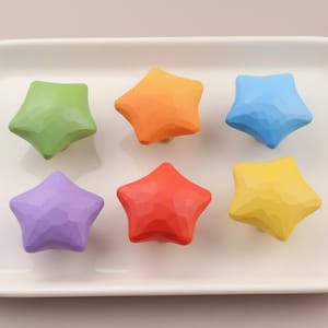May include: Six colorful star-shaped decorative objects in green, orange, blue, purple, red, and yellow are arranged on a white rectangular tray. The stars have a textured surface and are evenly spaced.