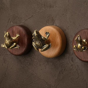 May include: Three decorative wall hooks featuring brass frog figurines mounted on wooden discs. The discs are in varying shades of brown and tan, creating a rustic and whimsical aesthetic. The hooks are ideal for hanging coats, keys, or other small items.