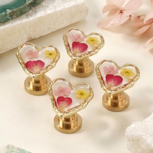 May include: Four heart-shaped cabinet knobs with a gold-toned metal base and a clear resin top. Each knob contains embedded dried flowers, including small pink and yellow blossoms and a single red petal, arranged within the heart shape.
