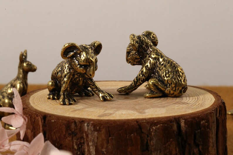 K&ouml;nnte beinhalten: Zwei Koala-B&auml;ren-Figuren aus Bronze werden auf einer Holzscheibe pr&auml;sentiert, mit einer K&auml;nguru-Figur im Hintergrund. Die Koalas posieren in einer spielerischen Interaktion. Die Figuren haben eine detaillierte, strukturierte Oberfl&auml;che.