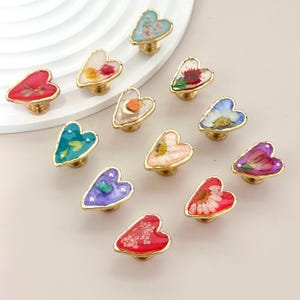 May include: A collection of heart-shaped cabinet knobs with gold-colored bases. Each knob features a different color and type of dried flower encased in clear resin. The knobs are arranged on a light-colored surface.