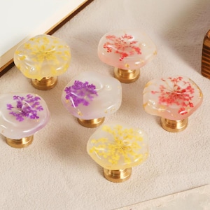 May include: Six decorative cabinet knobs with brass bases. Each knob features a clear, rounded top with dried flowers in shades of yellow, pink, and purple. The knobs are arranged on a light-colored surface.