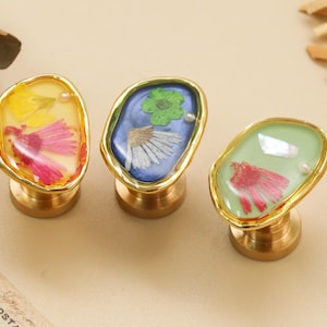 Può includere: Tre pomelli decorativi per mobili con basi in ottone e cornici irregolari color oro. Ogni pomello presenta una resina di colore diverso con fiori secchi e una piccola perla. I colori della resina sono giallo, blu e verde.
