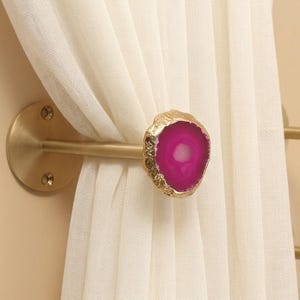 May include: A wall-mounted curtain tieback featuring a circular gold-toned metal base and rod, finished with a polished, vibrant pink agate slice at the end. The natural stone has a textured, gold-colored metallic edge and holds a sheer white curtain.