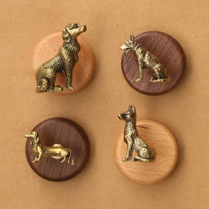 May include: Four circular cabinet knobs featuring brass-toned metal dog figurines mounted on round wooden bases. Two knobs have light-colored wood bases, and two have dark-colored wood bases. The dog figures include a sitting retriever, a sitting shepherd, a standing dachshund, and a sitting shepherd.