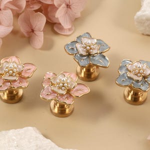 Könnte beinhalten: Vier dekorative Schrankknöpfe in Blumenform. Zwei sind rosa und weiß, und zwei sind blau und weiß, alle mit goldfarbenen Basen und perlenartigen Zentren. Die Knöpfe sind auf einer hellbraunen Oberfläche mit floralen Akzenten angeordnet.