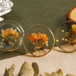 May include: Three clear resin pendants with dried orange flowers and small white beads. Each pendant has a gold-colored rim. The pendants are displayed on a neutral surface with a silver decorative element and a wooden slice in the background.