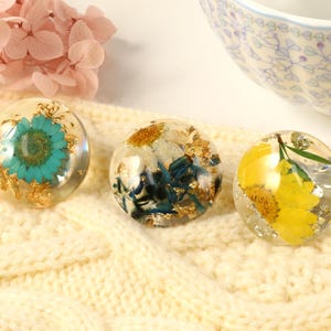 Może przedstawiać: Trzy okrągłe ozdoby z żywicy z prasowanymi kwiatami i złotem. Ozdoby przedstawiają niebieski kwiat, biały kwiat i żółty kwiat.