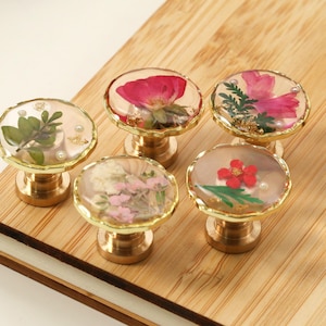 May include: A collection of decorative cabinet knobs. Each knob features a brass base and a clear resin top, encasing dried flowers. The knobs are displayed on a wooden surface, showcasing their unique floral designs.