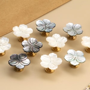 Puede incluir: Un conjunto de tiradores de armario con forma de flor. Los tiradores presentan un diseño de flor de nácar en blanco y gris, montados sobre una base dorada. Los tiradores están dispuestos sobre una superficie beige claro.