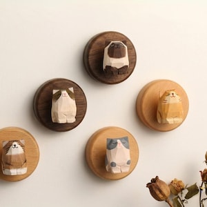 May include: A collection of six wooden cat figurines mounted on circular wooden bases. The cats are carved in a geometric style, with varying colors and patterns. The bases are affixed to a white wall.
