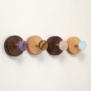 May include: Four decorative wall hooks with wooden bases and gemstone knobs. The hooks feature amethyst, tiger's eye, rose quartz, and opal knobs. The wooden bases are in varying shades of brown and natural wood tones, with brass hardware.