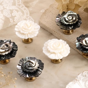 May include: Six decorative floral cabinet knobs. Three are white, and three are black and grey with a pearlescent sheen. Each knob has a brass base and is shaped like a rose.