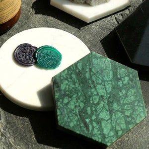 May include: Three wax seals on a white marble surface. One seal is green with a crescent moon and star design. The other two seals are purple with a floral design. The seals are on a white marble surface next to a green hexagonal marble coaster.