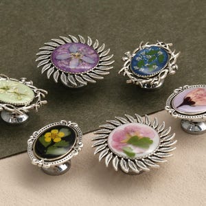 May include: A selection of decorative cabinet knobs with pressed flower designs. Each knob has a different flower encased in resin, set in a silver-coloured metal base. The flowers are various colours, including purple, pink, yellow, and green.