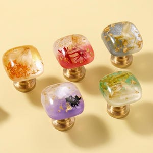May include: Five decorative cabinet knobs with square, translucent tops in various colours, including orange, red, purple, green, and grey. Each knob features gold-coloured accents and a brass base. The knobs are arranged on a light yellow surface.