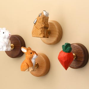 Könnte beinhalten: Ein Set aus vier Wandhaken mit handgeschnitzten Holzfiguren, darunter zwei Hasen, ein braunes Tier und eine orangefarbene Karotte mit grünen Blättern. Jede Figur ist auf einem runden Sockel aus hellem oder dunklem Holz montiert.