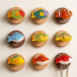 May include: A set of nine wall-mounted hooks, each featuring a hand-carved wooden fish figure painted in various colors including orange, yellow, blue, and green. The fish are mounted on circular wooden bases in light and dark wood tones.