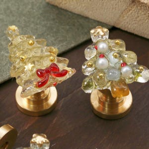 May include: Two Christmas tree-shaped wax seal stamps with brass bases. One has a red bow and gold accents, the other has white and red accents. The trees are made of clear, textured material.