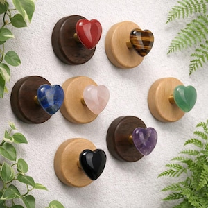 Può includere: Una collezione di ganci a muro a forma di cuore in pietra preziosa. I ganci presentano varie pietre preziose, tra cui rosso, occhio di tigre, blu, rosa, verde, nero e viola, montate su basi di legno. I ganci sono montati su una parete bianca.