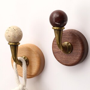 Op de afbeelding: Twee wandhaken met ronde houten voetjes, een lichtgekleurde en een donkergekleurde. Elke haak heeft een gebogen messingkleurige metalen arm met een bolvormige knop, een van gestructureerde beige steen en een van gladde bruine steen.