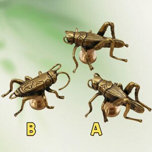 May include: Three bronze-coloured cricket-shaped decorative objects. Each cricket has detailed legs, antennae, and body segments. The objects are mounted on a round, gold-toned base. Letters A and B are in the image.