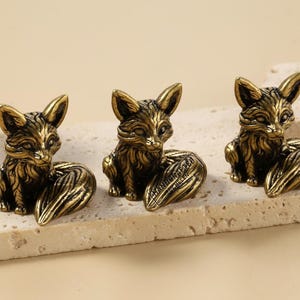 Könnte beinhalten: Drei bronzefarbene Fuchsstatuen mit detailliertem Fell und eingerollten Schwänzen. Die Füchse sitzen aufrecht, die Ohren aufgestellt. Die Figuren werden auf einer hellen, strukturierten Oberfläche präsentiert.