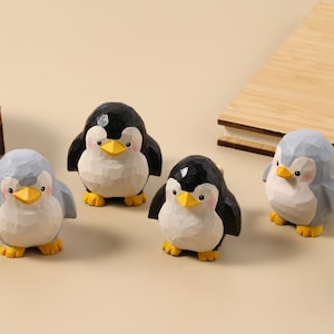 Könnte beinhalten: Vier Holzpinguin-Figuren mit schwarzen, weißen und grauen Körpern, gelben Schnäbeln und Füßen. Die Pinguine sind auf einer hellbeige Oberfläche angeordnet. Die Figuren haben ein geschnitztes Aussehen, was auf handwerkliche Herstellung hindeutet.