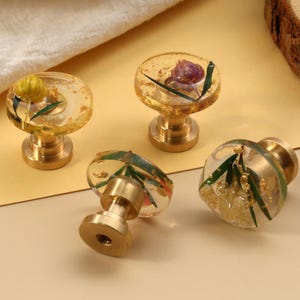 May include: Four decorative cabinet knobs with clear resin tops, each containing botanical elements and gold flakes. The knobs have brass bases and are arranged on a light yellow surface, showcasing a unique design.