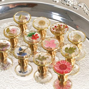 Puede incluir: Una colección de tapones decorativos para botellas con bases doradas y tapas transparentes y redondeadas. Cada tapón presenta una flor seca o elemento botánico diferente encapsulado en resina. Los tapones están dispuestos sobre una bandeja plateada.