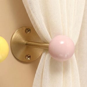 Könnte beinhalten: Ein an der Wand montierter Vorhanghalter mit einer runden Basis aus gebürstetem Messing mit zwei Schraublöchern, einer horizontalen Messingstange und einem glatten, glänzenden, blassrosa kugelförmigen Knopf, der einen cremeweißen Vorhang hält.