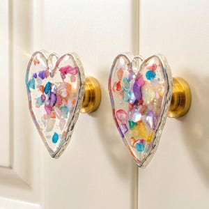 Op de afbeelding: Twee hartvormige kastknoppen met een voorkant van heldere hars gevuld met veelkleurige verbrijzelde steenfragmenten in roze, blauw, paars en geel. Elke knop heeft een goudkleurige cilindrische basis en een zilverkleurige rand.