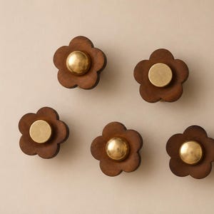 Op de afbeelding: Vijf bloemvormige kastknoppen. Elke knop heeft een donkerbruine houten bloem met vijf bloemblaadjes en een ronde, goudkleurige kern. De knoppen zijn geplaatst tegen een neutrale achtergrond.