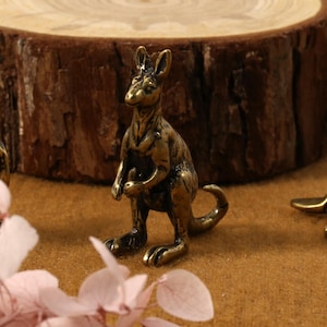 K&ouml;nnte beinhalten: Bronze-K&auml;nguru-Figur, die aufrecht steht, mit einem Beutel und einem eingerollten Schwanz. Weitere Bronze-Tierfiguren sind sichtbar, darunter ein Koala und ein Schnabeltier. Die Figuren stehen auf einer braunen Oberfl&auml;che, mit einer Holzscheibe im Hintergrund.