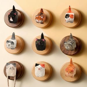 May include: Nine wooden cat-shaped wall hooks in various colours and patterns. Each cat is mounted on a round wooden base. The cats are black, orange, white, and grey, with details like stripes and spots. Some have tails up.