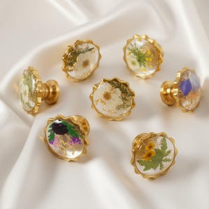 Puede incluir: Un conjunto de tiradores dorados para armarios con caras de resina transparente, cada uno con flores secas y elementos botánicos. Los tiradores están dispuestos sobre una superficie de satén blanco, mostrando sus diseños florales únicos y acentos dorados.