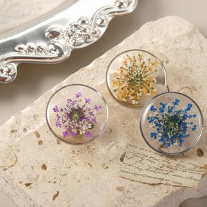 Könnte beinhalten: Drei klare, runde Harzuntersetzer mit getrockneten Blumen in Lila, Gelb und Blau. Jeder Untersetzer hat einen silbernen Metallrand. Die Untersetzer werden auf einer strukturierten, hellfarbigen Steinoberfläche präsentiert.