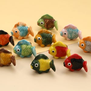 Może przedstawiać: Kolekcja jedenastu ceramicznych figurek ryb w różnych kolorach. Każda ryba ma unikalny wzór, z kolorami takimi jak pomarańczowy, niebieski, zielony, żółty i czerwony. Ryby są ułożone na jasnożółtej powierzchni.