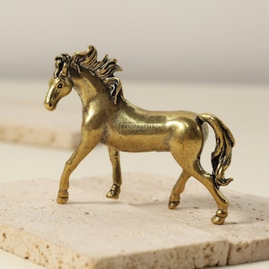 Peut inclure: Figurine de cheval en laiton doré, avec une crinière et une queue détaillées. Le cheval est en position de marche, les pattes tendues. La figurine est posée sur une surface texturée et claire. Le texte "HANDCRAFTED by jiten" est visible.