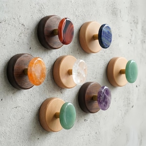 May include: A collection of decorative wall hooks with wooden bases and gemstone knobs. The hooks feature various gemstones, including red agate, lapis lazuli, clear quartz, green jade, and amethyst. The wooden bases are in shades of light and dark brown.