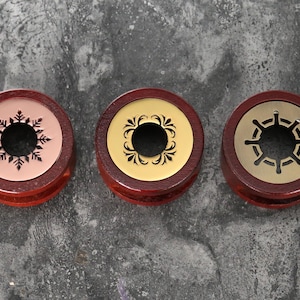 Pode incluir: Três medidores de orelha de madeira com diferentes designs. O primeiro medidor tem um fundo rosa com um design de floco de neve preto. O segundo medidor tem um fundo dourado com um design floral preto. O terceiro medidor tem um fundo dourado com um design de roda de navio preto.