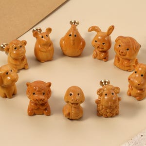 May include: A collection of ten wooden animal figurines, each with a small gold-colored knob on top. The figurines include a monkey, dog, tiger, sheep, cow, pig, rabbit, and a duck. The wood has a warm, natural tone.
