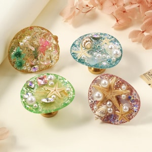 May include: Four decorative cabinet knobs with ocean-themed designs. Each knob features a resin dome with embedded seashells, starfish, and faux pearls. The knobs have brass-coloured bases and are in shades of green, blue, and brown.