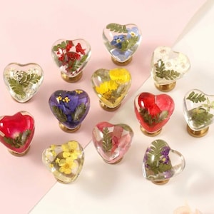 Op de afbeelding: Een verzameling van twaalf hartvormige kastknoppen van heldere hars, elk met geperste gedroogde bloemen en groen in verschillende kleuren, waaronder rood, geel, blauw, wit en groen, bevestigd op kleine ronde metalen voetjes.