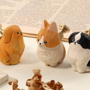 Könnte beinhalten: Drei Holzfiguren von Hunden mit Messingaufsätzen. Die Hunde sind in den Farben Beige, Weiß und Beige sowie Schwarz und Weiß gehalten. Der mittlere Hund hat ein Etikett mit der Aufschrift "HANDCRAFTED BY HELEN".