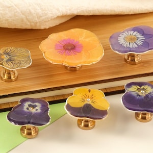 May include: Six decorative cabinet knobs with flower designs. The knobs feature pressed flowers in shades of yellow, orange, and purple, set in clear resin. Each knob has a gold-coloured base and a unique floral pattern.