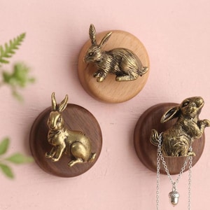 May include: Three wall-mounted hooks featuring brass-toned rabbit figures attached to circular wooden bases. Two hooks are made of dark wood and one is made of light wood. A silver-toned chain necklace with an acorn pendant hangs from one rabbit.