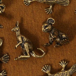 K&ouml;nnte beinhalten: Eine Sammlung kleiner, bronzefarbener Tierfiguren, darunter ein K&auml;nguru, ein Koala und ein Schnabeltier, sind auf einer braunen Oberfl&auml;che angeordnet. Die detaillierten Figuren haben eine Vintage-&Auml;sthetik mit sichtbaren Texturen und Formen.