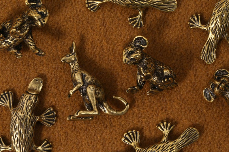 K&ouml;nnte beinhalten: Eine Sammlung kleiner, bronzefarbener Tierfiguren, darunter ein K&auml;nguru, ein Koala und ein Schnabeltier, sind auf einer braunen Oberfl&auml;che angeordnet. Die detaillierten Figuren haben eine Vintage-&Auml;sthetik mit sichtbaren Texturen und Formen.