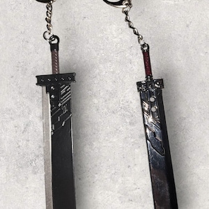 May include: Two sword-shaped keychains. The blades are dark gray with silver accents, and the handles are brown and red. Each keychain has a silver chain and a ring for attachment. The keychains are displayed against a gray background.